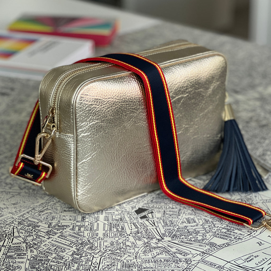 Large leather gold crossbody bag with blue, yellow and red striped slim interchangeable strap and navy blue large tassel 
