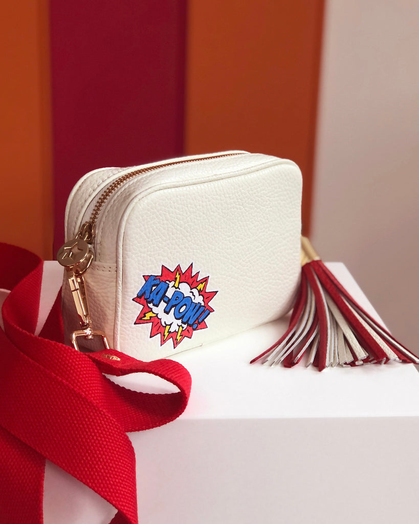 Small white leather cross body bag with graphic painting phrase 'KA-POW' in both left corner with red slim fabric interchangeable strap.